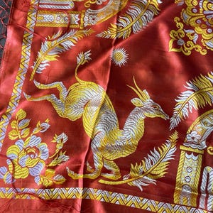 May include: A red textile with intricate gold and silver embroidery. The design features a leaping deer, floral patterns, and ornate borders. The fabric has a silky sheen and a fringed edge, showcasing a detailed, artistic style.