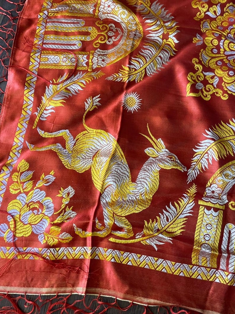 May include: A red textile with intricate gold and silver embroidery. The design features a leaping deer, floral patterns, and ornate borders. The fabric has a silky sheen and a fringed edge, showcasing a detailed, artistic style.