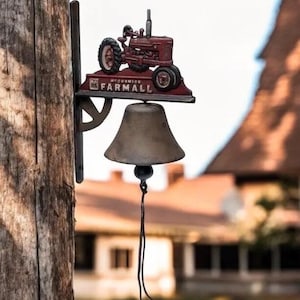 Può includere: Una campana decorativa con un design di trattore rosso e la scritta "McCormick Farmall". La campana è montata su un palo di legno, con un'estetica rustica. La campana ha un cordino di trazione marrone scuro.