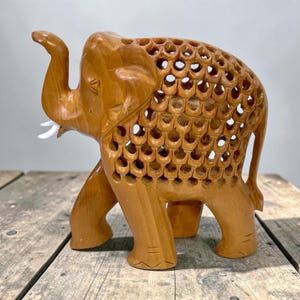 May include: A wooden elephant sculpture with intricate openwork detailing on its back. The elephant is carved from light brown wood, with white tusks and a raised trunk. The sculpture is displayed on a rustic wooden surface.