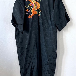 May include: Black kimono-style robe with an embroidered orange and gold dragon design on the back. The robe has short sleeves and a floral pattern. The dragon is detailed with green and pink accents.