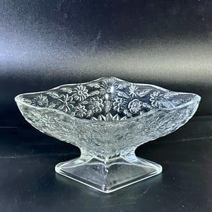 Könnte beinhalten: Klarer Glas-Servierteller mit floralem Muster. Die Schale hat eine rautenförmige Form mit einem Sockel. Das florale Design ist in das Glas geätzt. Der Sockel ist quadratisch. Der Hintergrund ist schwarz.