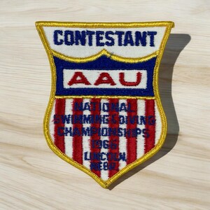 May include: A vintage shield-shaped patch with gold trim. The top section reads "CONTESTANT" in blue. Below, "AAU" is in red on a blue background. The bottom section has "NATIONAL SWIMMING & DIVING CHAMPIONSHIPS 1968 LINCOLN NEB".