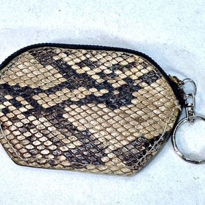 Pode incluir: Uma pequena carteira de moedas com um padrão de pele de cobra em tons de bege, castanho e preto. A carteira tem um fecho de correr preto e um porta-chaves prateado. A forma geral é um hexágono arredondado.