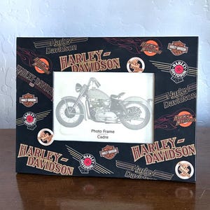 May include: A rectangular black photo frame featuring a repeating Harley-Davidson logo pattern in white, orange, and red. The frame includes various brand emblems and flame graphics. A central white insert displays a motorcycle illustration and text.