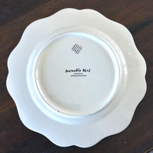 Op de afbeelding: Een witte keramische plaat met een geschulpte rand. De plaat heeft de tekst "Nathalie Lete" en "exclusively for Anthropologie" op de achterkant gedrukt. De plaat ligt op een donkerbruin oppervlak.