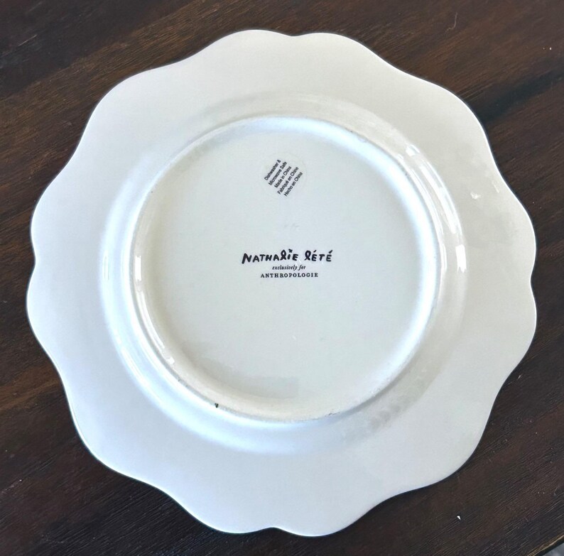 Op de afbeelding: Een witte keramische plaat met een geschulpte rand. De plaat heeft de tekst "Nathalie Lete" en "exclusively for Anthropologie" op de achterkant gedrukt. De plaat ligt op een donkerbruin oppervlak.