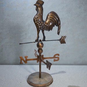 May include: A bronze-toned metal weather vane featuring a rooster figure standing atop a horizontal arrow. Below the arrow, a central rod supports a directional cross with the letters N, S, E, and W, all mounted on a circular base.