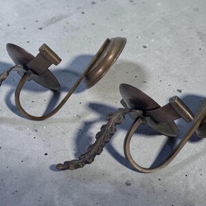 May include: Two bronze-toned earrings. Each earring features a circular disc, a rectangular bar, and a curved, open ring design. One earring has a twisted stem, while the other has a leaf-like stem.