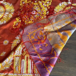 May include: A detailed shot of a silk scarf with intricate patterns. The scarf features a rich red base with gold, white, and yellow floral and architectural designs. The border is a mix of purple and gold, with red fringe.