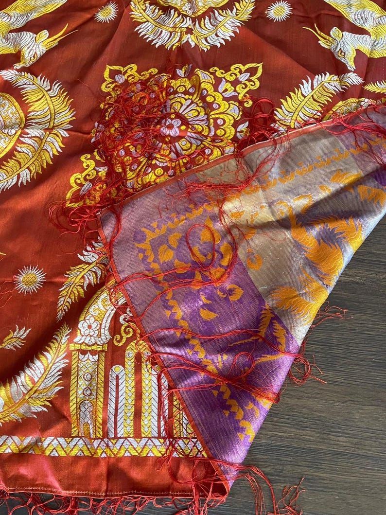 May include: A detailed shot of a silk scarf with intricate patterns. The scarf features a rich red base with gold, white, and yellow floral and architectural designs. The border is a mix of purple and gold, with red fringe.
