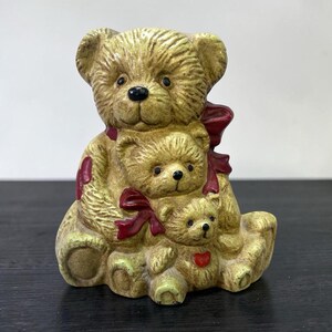 May include: A tan ceramic figurine featuring three stacked teddy bears with textured fur. The bears are adorned with dark red bows and a small red heart on the bottom bear. Each bear has black eyes and a black nose, creating a cohesive sculpture.
