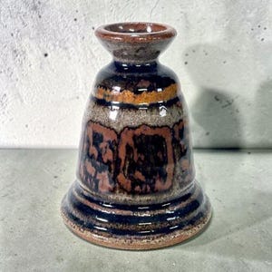 May include: A bell-shaped ceramic candle holder featuring a glossy glaze with horizontal bands of dark brown, black, and tan. The piece has a wide, flared base, a tapered neck, and a circular opening at the top with a smooth, reflective finish.