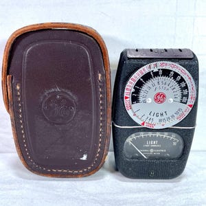 May include: A vintage General Electric light meter with a brown leather case. The meter is black with a circular dial and a needle gauge. The dial has numbers and markings for light measurement.