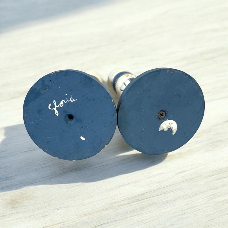 May include: Two wooden candle holders with navy blue bases and white and blue striped bodies. The word "Gloria" is handwritten on the bottom of one base. The other base has a white crescent moon design.