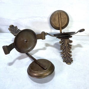 May include: Two antique brass candle holders. One has a circular base and a pivoting candle cup. The other has a decorative leaf design and a small candle cup. Both are made of brass and have a vintage look.