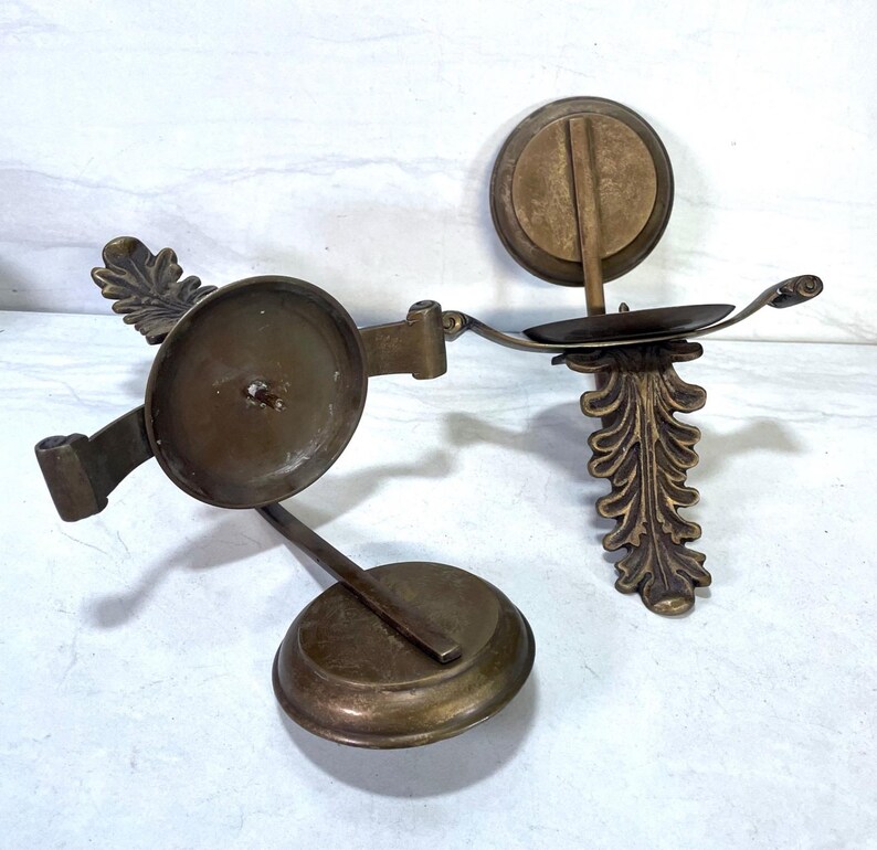 May include: Two antique brass candle holders. One has a circular base and a pivoting candle cup. The other has a decorative leaf design and a small candle cup. Both are made of brass and have a vintage look.