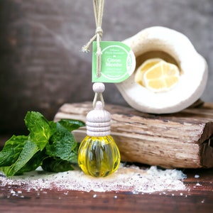 May include: A small glass bottle with a wooden cap and a yellow liquid inside. The bottle is hanging from a string and is surrounded by green mint leaves and a white ceramic bowl with a lemon slice inside. The bottle has a label that reads "Diffuseur d'Huile Essentielle Citron Menthe Mint Lemon".