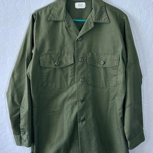 May include: An olive green long-sleeve button-up shirt featuring a pointed collar, two chest pockets with buttoned flaps, and a central button placket. A white rectangular label at the inner neckline displays the text 15 1/2 33.