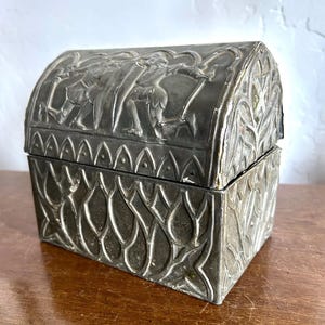 May include: A rectangular metal chest with a hinged, arched lid. The exterior features embossed relief designs, including figures of knights with shields and swords on the lid, and a repeating pointed leaf or arch pattern on the base.