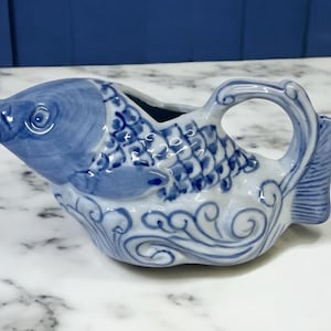 May include: A blue and white ceramic creamer in the shape of a fish. The fish has detailed scales, fins, and a handle. The creamer features a blue and white color scheme with a decorative wave pattern.