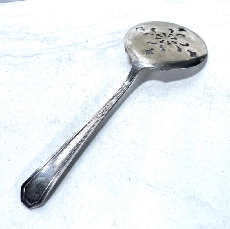May include: A silver-toned slotted spoon with a decorative floral design on the head. The handle is long and slender, with a geometric end. The text "A Silver Plate" is visible on the handle. This kitchen utensil is likely used for serving.