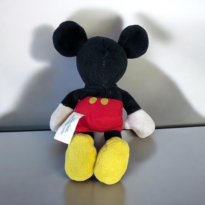 Puede incluir: Un peque&ntilde;o peluche de Mickey Mouse. El juguete tiene cabeza y orejas negras, pantalones cortos rojos con dos botones amarillos, pies amarillos y guantes blancos. Una etiqueta blanca est&aacute; adherida al juguete.