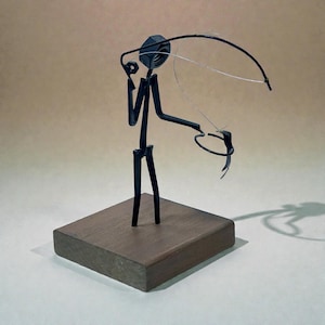 May include: A metal sculpture of a figure fishing, constructed from dark steel rods and a hexagonal nut for a head. The figure holds a curved wire rod with a thin fishing line attached to a small metal fish, all mounted on a square wooden base.