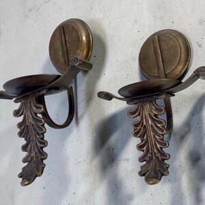 May include: Two antique bronze wall sconces. Each sconce features a circular backplate with a vertical groove, a curved arm holding a small dish, and a decorative leaf design. The sconces are mounted on a light gray wall.