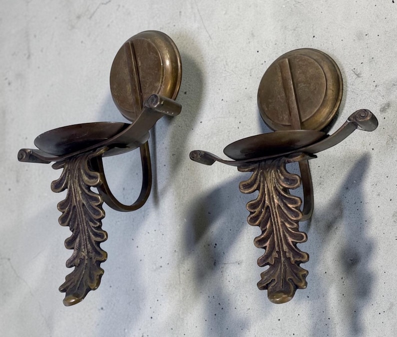 May include: Two antique bronze wall sconces. Each sconce features a circular backplate with a vertical groove, a curved arm holding a small dish, and a decorative leaf design. The sconces are mounted on a light gray wall.