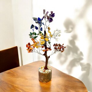 May include: A decorative tree sculpture with a brown trunk and branches made of silver wire. The leaves are colorful gemstones in shades of red, orange, yellow, green, blue, and purple. The tree sits in a wooden base.