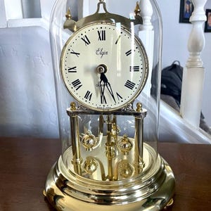 May include: A gold-toned Elgin quartz clock with a glass dome. The clock face is cream-colored with black Roman numerals and hands. The base and decorative elements are gold, and the clock has a pendulum.
