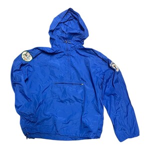 May include: Bright blue hooded anorak jacket with a front pocket and a zipper closure. The jacket features two patches on the sleeves. The jacket is made of a lightweight, water-resistant material.