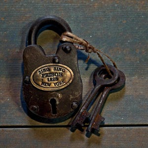 May include: A dark, weathered metal padlock with a brass oval plate featuring the text SING SING PRISON 1825 NEW YORK. Two matching skeleton keys are attached to the shackle with a thin, frayed piece of twine, showing a rustic, aged finish.