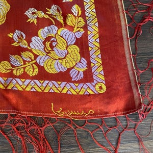 May include: A red textile with floral embroidery in yellow and lavender. The design includes a geometric border and fringe. The textile has a gold trim and an embroidered signature in gold thread.