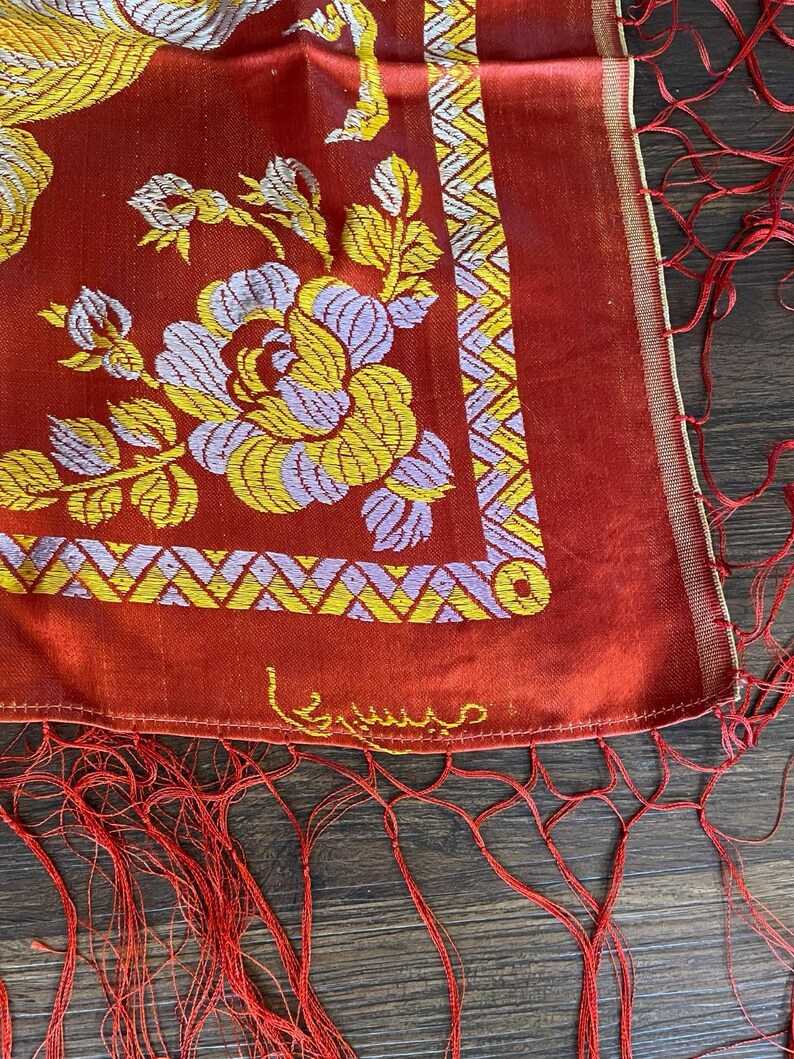 May include: A red textile with floral embroidery in yellow and lavender. The design includes a geometric border and fringe. The textile has a gold trim and an embroidered signature in gold thread.