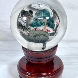 May include: A clear glass sphere with an Asian-inspired landscape painting inside, set on a dark wooden stand. The painting features a pagoda, trees, and mountains in shades of gray, green, and red. The stand is a rich, polished brown.