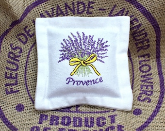May include: White fabric sachet with lavender embroidery and the word "Provence". The sachet is sitting on a burlap bag with purple text that reads "FLEURS DE LAVANDE - LAVENDER FLOWERS - PRODUCT OF FRANCE".