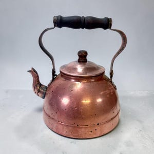 Op de afbeelding: Een koperen ketel met een donkerhouten handvat en knop. De ketel heeft een gebogen tuit en een afgeronde body. Het metaal heeft een warme, roodbruine tint, met enkele donkere vlekken. De handgreep is bevestigd met gebogen metalen steunen.
