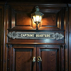 Pode incluir: Porta de madeira escura com uma placa dourada que diz "CAPTAINS QUARTERS". Acima, uma lanterna vintage iluminada. A porta tem puxadores de latão e faz parte de uma parede com painéis de madeira.