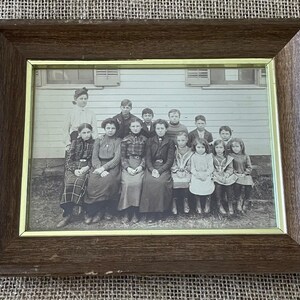 May include: A framed black and white photograph of a group of people, likely a class or family portrait. The frame is dark brown with a gold inner border. The photograph shows a group of people in front of a building.