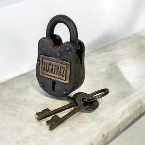 May include: A weathered, dark metal padlock with the word "ALCATRAZ" on a rectangular plate. Two antique keys are placed near the lock. The lock features a curved handle and appears to be made of iron or steel.