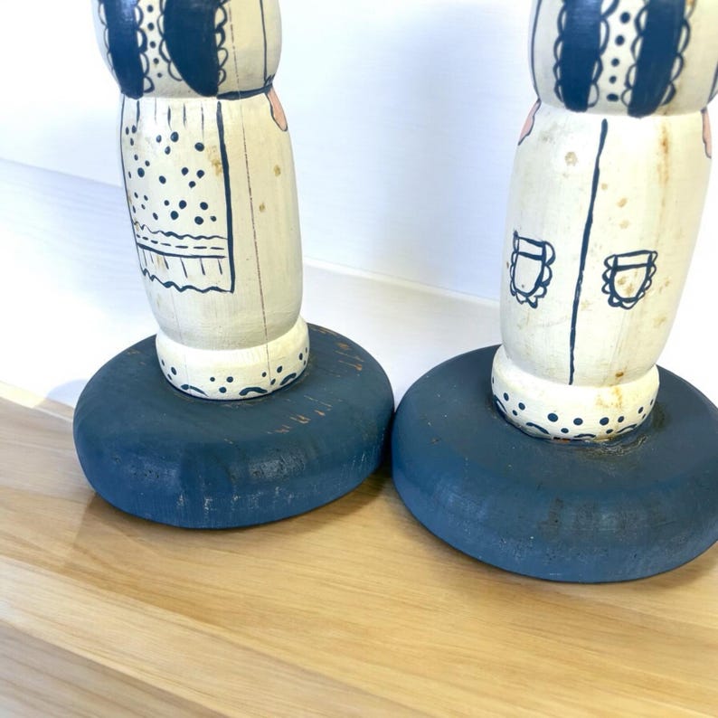 May include: Two wooden decorative figures with a cream-colored body and blue accents. The figures have a round, dark blue base. The upper portion of the figures have painted details, including a bib-like design and decorative patterns.