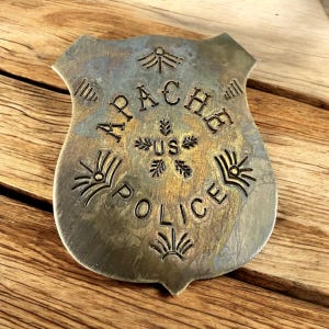 May include: A vintage silver-toned Apache Police badge. The badge is shield-shaped with the words "APACHE US POLICE" engraved in a bold, serif font. Decorative sunburst and geometric designs surround the text. The badge has a tarnished, aged appearance.