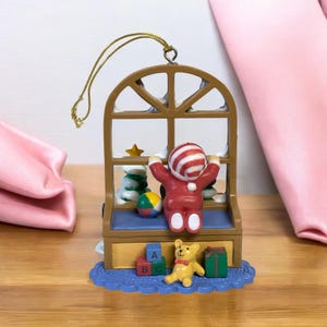 May include: A Christmas ornament featuring a child in a red and white striped hat and suit, reaching up towards a brown window. The ornament includes a teddy bear, ABC blocks, a ball, and a wrapped gift. The ornament is on a blue scalloped base.