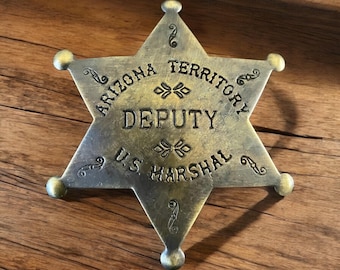 Könnte beinhalten: Ein sechszackiger Sternabzeichen im Vintage-Stil mit den Worten "ARIZONA TERRITORY DEPUTY U.S. MARSHAL" in erhabenen Buchstaben. Das Abzeichen hat eine metallische, antike Goldoberfläche und dekorative Verzierungen.