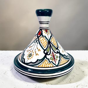 May include: A white, cone-shaped ceramic tagine pot with a dark green base and lid. The pot features a detailed floral pattern with yellow, red, and green accents. The lid has a cylindrical handle with white and dark green stripes.
