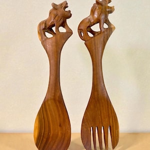 Puede incluir: Un par de utensilios para ensalada de madera. Uno es una cuchara, el otro un tenedor. Cada mango está rematado con un león de madera tallada. Los utensilios son de madera marrón claro con un acabado liso.
