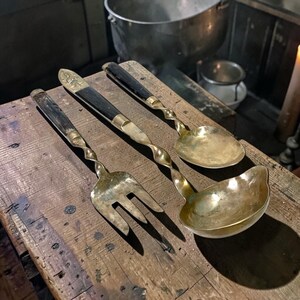 May include: Antique brass serving utensils set on a wooden surface. The set includes a fork, a knife, and two spoons. The handles are black and the utensils have a twisted design. The fork is 10 inches long.