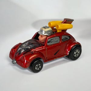 May include: A vintage red toy car with a silver helmeted driver and yellow exhaust pipes. The car has black wheels and a black and silver design on the hood. The toy car is a classic collectible.